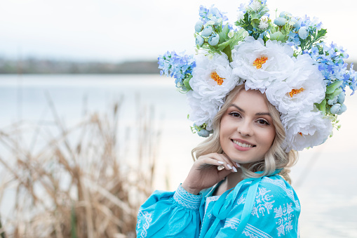 Beautiful young Ukrainian woman dressed in a blue embroidered dress with a large beautiful floral wreath on her head smiling, looking at the camera.