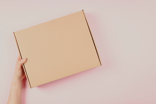 Top view to female hand holding brown cardboard box on pastel pink background. Mockup parcel box. Packaging, shopping, delivery concept.
