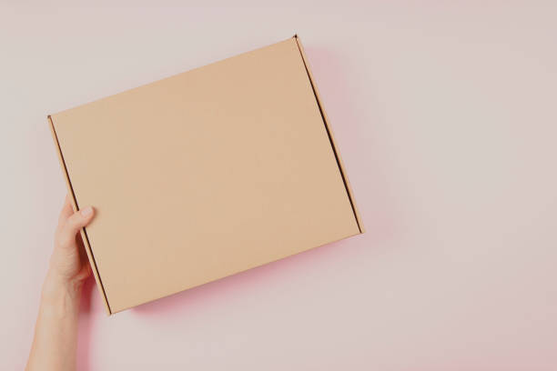 Female hand holding brown cardboard box on pastel pink background. Top view to mockup parcel box. Packaging, shopping, delivery concept stock photo