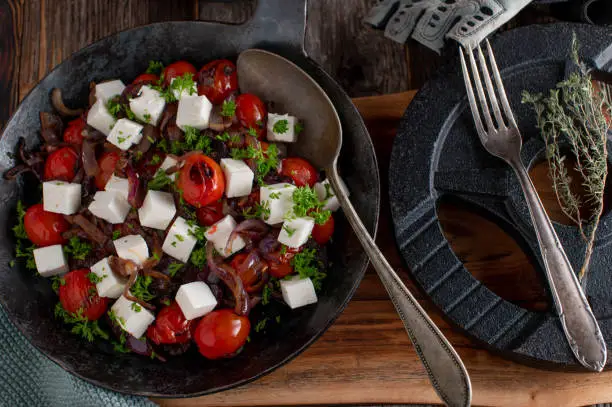 Fitness meal for dinner or lunch with roasted tomatoes and red onions. Marinated with olive oil and topped with real sheep cheese. Warm salad. Served with dumbbell on wooden table background.