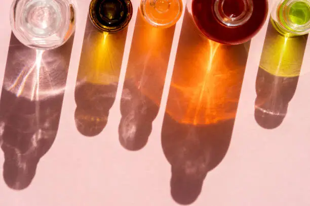 Different cosmetic glass bottles standing on pink background. Abstract pattern by shadows and illuminating reflections in the sunlight. Red, green and yellow shades. Flat lay style with copy space