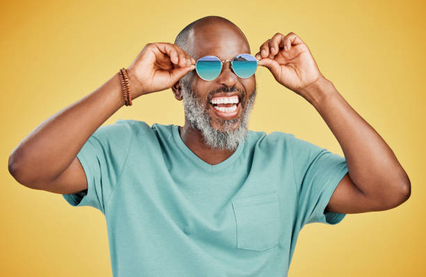 Happy mature African American man standing alone against yellow background in a studio and posing with sunglasses. Smiling black man feeling fashionable and cool while wearing glasses. Summer and beach Happy mature African American man standing alone against yellow background in a studio and posing with sunglasses. Smiling black man feeling fashionable and cool while wearing glasses. Summer and beach fashion model men male sunglasses stock pictures, royalty-free photos & images