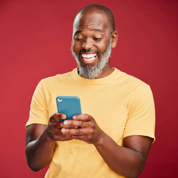 un hombre afroamericano feliz de pie sobre un fondo rojo de estudio, sosteniendo y usando su teléfono celular para navegar por internet. hombre negro sonriente riendo mientras navega por su teléfono en busca de redes sociales - portrait laughing people men fotografías e imágenes de stock