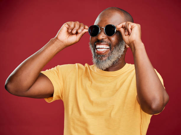 happy mature african american man standing alone against a red background in a studio and posing with sunglasses. smiling black man feeling fashionable and cool while wearing glasses. summer and beach - fashionable studio shot indoors lifestyles imagens e fotografias de stock