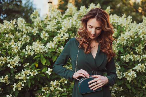 a young beautiful adult woman in a green dress stands against a background of flowering bushes and holds a small green purse in her hands.