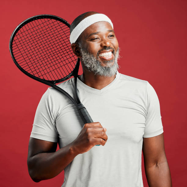 one happy mature african american man standing against a red background in studio and posing with a tennis racquet. smiling black man feeling fit and sporty while playing a match. ready for the court - tennis active seniors healthy lifestyle senior men imagens e fotografias de stock