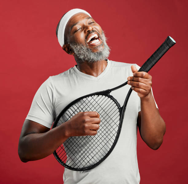 ein reifer afroamerikanischer mann, der einen tennisschläger als gitarre benutzt und vor einem roten studiohintergrund singt. schwarzafrikaner mit einem schweißband auf dem kopf, spaß und lächelnd beim singen. du musst deine stimme hörbar machen - tennis active seniors healthy lifestyle senior men stock-fotos und bilder