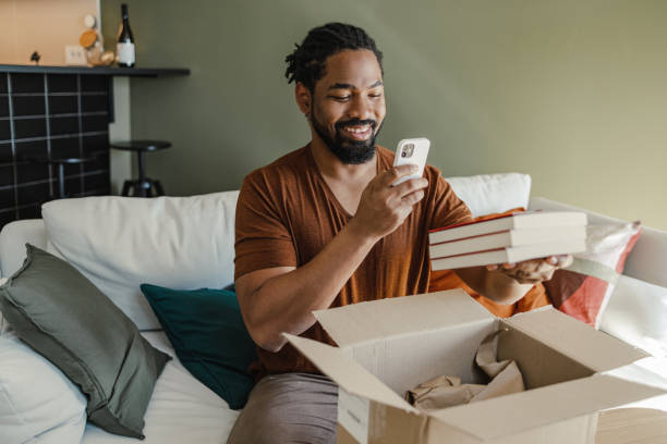 Young man unpacking the box and taking a picture of online ordered items Young African American man is at home. He is unpacking the delivered package and taking a picture of the ordered item. book bookstore sale shopping stock pictures, royalty-free photos & images