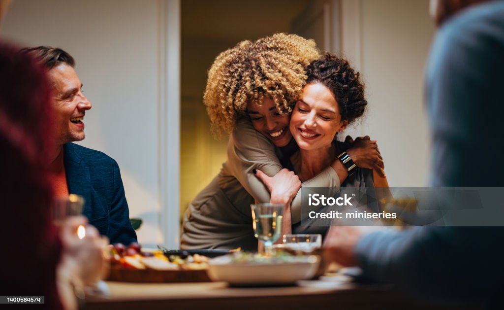 Two Friends Hugging During A Dinner Celebration Cheerful smiling African-American woman embracing her female friend during a dinner with family and friends. Family Stock Photo