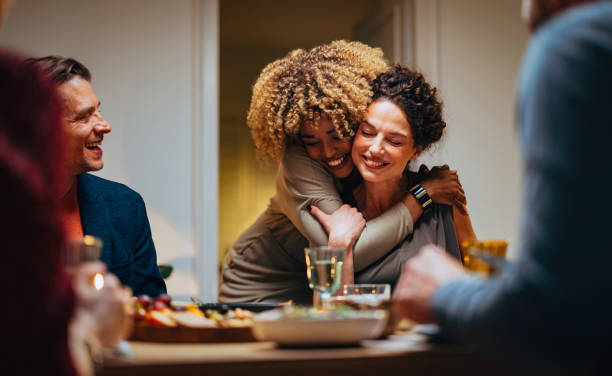 Two Friends Hugging During A Dinner Celebration Cheerful smiling African-American woman embracing her female friend during a dinner with family and friends. holidays and celebrations stock pictures, royalty-free photos & images