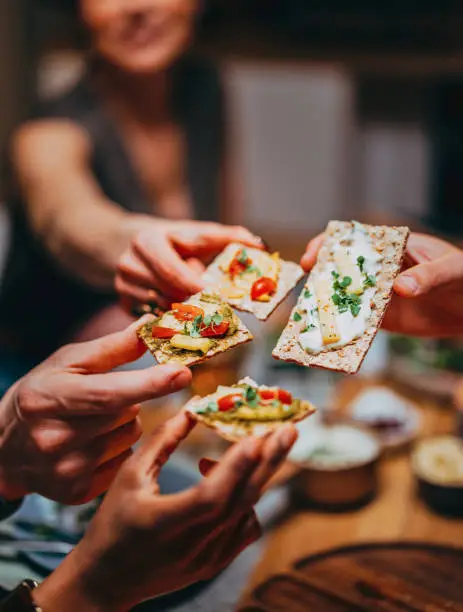 Close up photo of womans and mans hands holding crispbread with cream and cherry tomato at dining table.