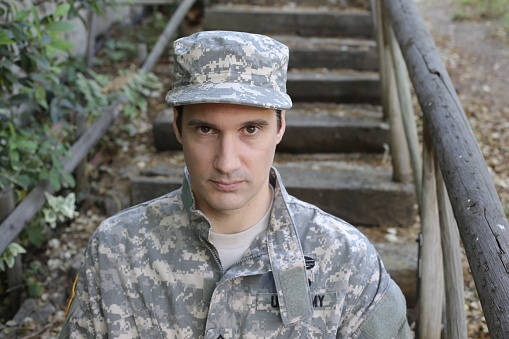 A caucasian army soldier in his 30s is  wearing a traditional USA army uniform with cap. He looks at camera and is outdoors.