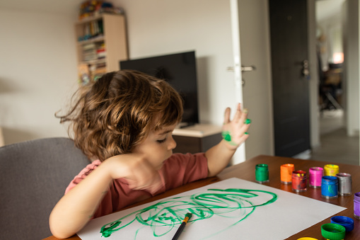Three year old cucasian boy with long hair playing with colors at home sitting at the table,painting with hands