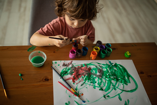 Three year old cucasian boy with long hair playing with colors at home sitting at the table,painting