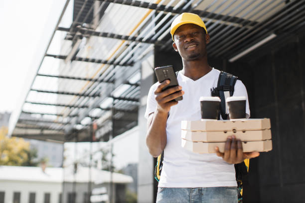 Courier of food delivery in yellow cap holding pizza boxes and takeaway coffee cups delivering order Courier of food delivery in yellow cap holding pizza boxes and takeaway coffee cups delivering order with thermal bag on his back. African courier with phone in hand looking address restaurant labor union stock pictures, royalty-free photos & images