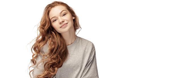 Happy redhead young girl with long curly hair looking at camera with light smile isolated on white background. Student in casual clothes. Concept of youth, beauty, life. Copy space for ad. Flyer