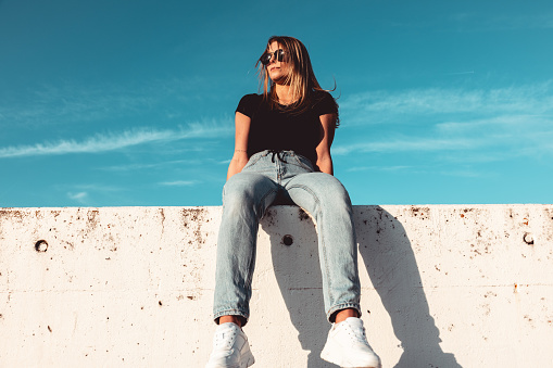 Happy Young woman wearing Black sunglasses sitting on modern concrete wall. Simplicity Modern Urban Style. Shot from below against the sky. Urban Lifestyle Youth Portrait.