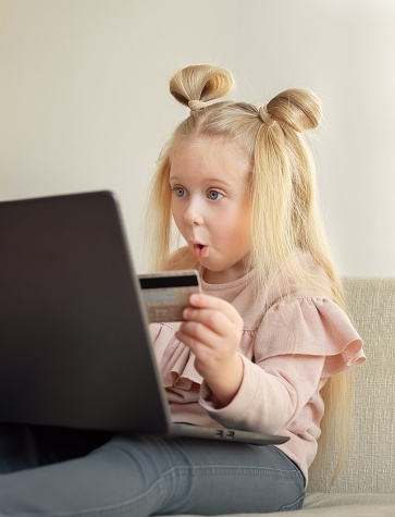 Funny blonde caucasian little girl holding credit card buying online at home