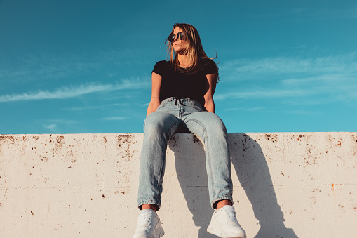 Young woman wearing Black sunglasses sitting on modern concrete wall. Simplicity Modern Urban Style. Shot from below against the sky. Urban Lifestyle Youth Portrait.
