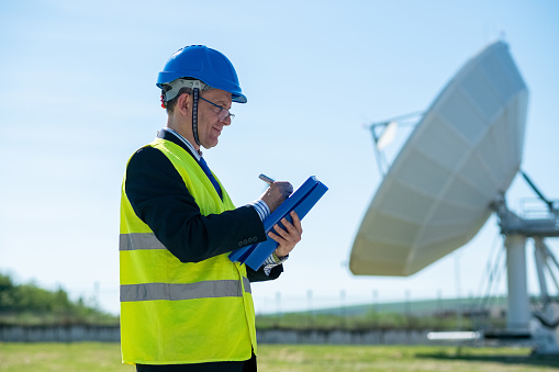 Telecom technician with helmet and reflective vest with blue folder and pen in his hands next to the satellite antenna of the telecom.