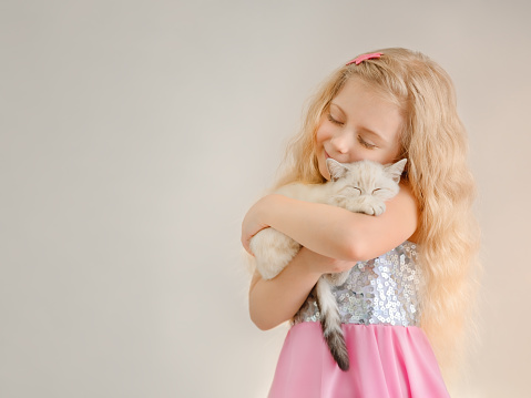 Little happy blonde girl  hugs a beige scottish sleeping kitten and smiles closing her eyes in a shiny dress.