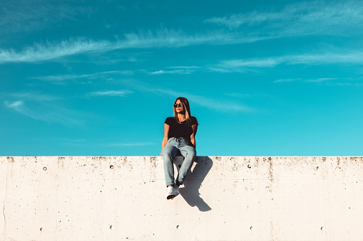 Young woman wearing large black sunglasses sitting on modern concrete wall. Simplicity Modern Urban Style. Shot from below against the sky. Urban Lifestyle Youth Portrait.