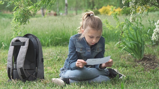 A pensive girl of nine years old, a schoolgirl, sits on the grass in the park. She holds a piece of paper in her hands and reads. High quality photo