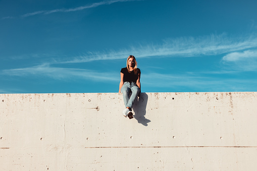 Young woman sitting on modern concrete wall. Simplicity Modern Urban Style. Shot from below against the sky. Urban Lifestyle Youth Portrait.