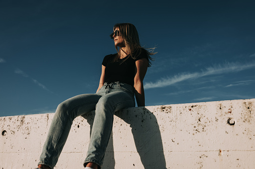 Young woman sitting on modern concrete wall with Sunglasses on. Simplicity Modern Urban Style. Shot from below against the sky. Urban Lifestyle Youth Portrait.
