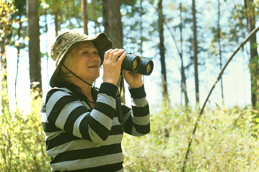Active smiling senior woman with binoculars in the forest. Traveling, hiking, walking, enjoying vacations