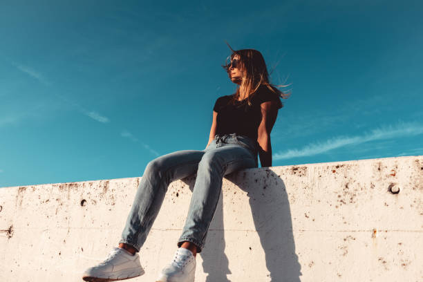 Cool Looking Young Woman Sitting on Concrete Wall With Sunglasses Fashion Portrait stock photo