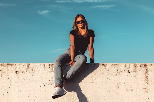 Young Woman Sitting on Concrete Wall With Sunglasses on. Simplicity Modern Urban Style. Shot from below against the sky. Urban Lifestyle Youth Portrait.