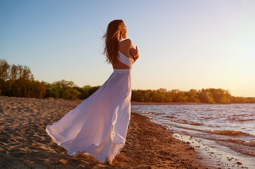 A beautiful young woman of Caucasian appearance with dyed hair posing on the seashore in the sun at sunset in a white dress in the wind. Romantic slender girl in nature enjoys relaxation