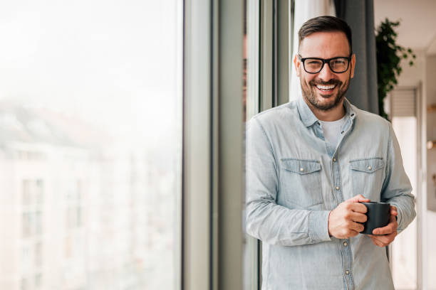 istock Smiling confident businessman having coffee while standing by window at office 1400610396