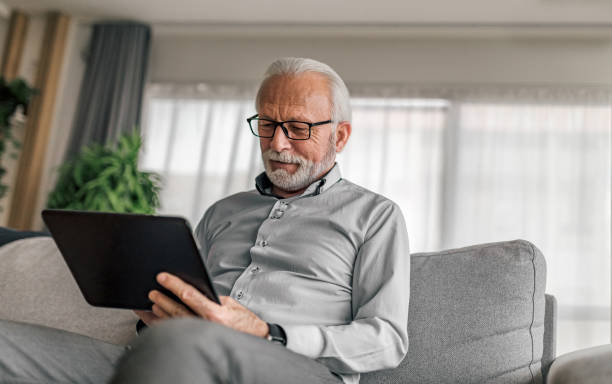 lächelnder älterer geschäftsmann mit digitalem tablet, während er zu hause auf der couch sitzt - senioren männer stock-fotos und bilder
