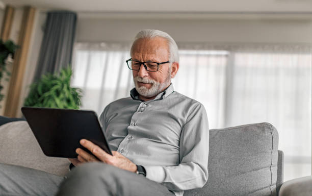 homme d’affaires âgé souriant utilisant une tablette numérique assis sur un canapé à la maison - hommes seniors photos et images de collection