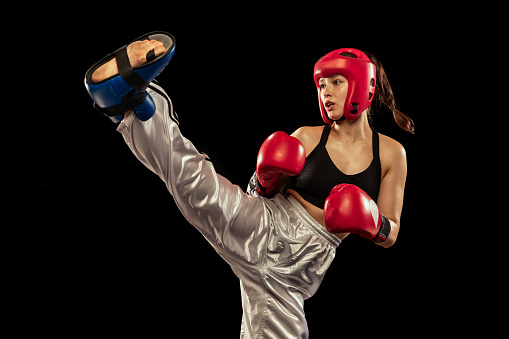 In action. Sportive woman, professional boxer in boxing gloves and helmet training isolated on dark studio background. Sport, competition, hobby, results, success concept. Achievements and challenges