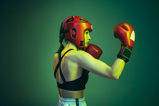 Peek-a-boo defense. Portrait of professional female boxer in sports protective equipment posing isolated on green background in neon. Sport, competition, hobby, results, success concept. Closeup