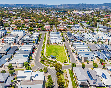 Aerial view of a prosperous, liveable city suburb, safe, connected, with open space park and trees, good roads, old and modern contemporary housing, multi-level, medium density housing and line of rolling hills in distance. Renewable energy solar panels on many building roofs. Shopping centre in distance.