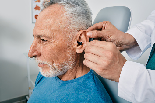 Grey-haired elderly man during installation intra-ear hearing aid into his ear by his doctor audiologist, close-up. Hearing treatment for hearing impaired people