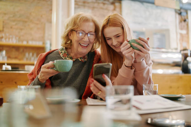 abuela feliz y su nieta adolescente sentadas en un café, disfrutando de un café y usando el teléfono inteligente para videos divertidos. - grandmother generation gap senior adult granddaughter fotografías e imágenes de stock