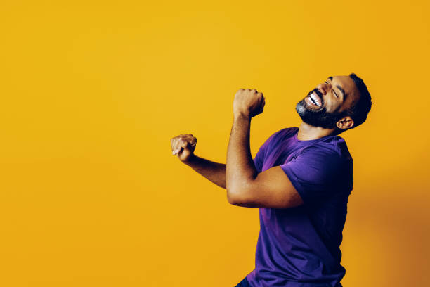 retrato de um homem vencedor de sucesso com uma barba e uma camiseta roxa celebrando dançando com o braço para cima em um fundo amarelo - arms raised behavior isolated one person - fotografias e filmes do acervo