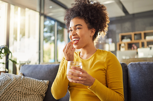Young happy mixed race woman taking medication with water at home. One hispanic female with a curly afro taking a vitamin for good health while sitting on the couch at home. Woman drinking a supplement