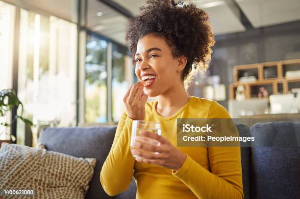 Young Happy Mixed Race Woman Taking Medication With Water At Home One Hispanic Female With A Curly Afro Taking A Vitamin For Good Health While Sitting On The Couch At Home Woman Drinking A Supplement Stock Photo - Download Image Now