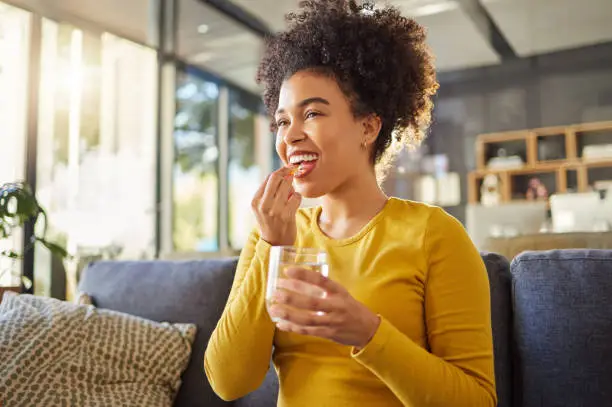 Photo of Young happy mixed race woman taking medication with water at home. One hispanic female with a curly afro taking a vitamin for good health while sitting on the couch at home. Woman drinking a supplement