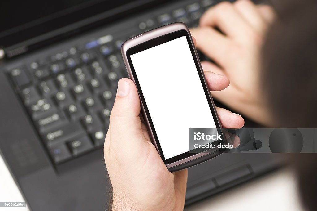 Hombre sosteniendo en blanco teléfono inteligente mientras usando una computadora portátil en la habitación - Foto de stock de Auricular de teléfono libre de derechos