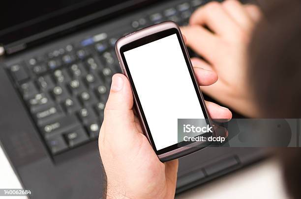 Hombre Sosteniendo En Blanco Teléfono Inteligente Mientras Usando Una Computadora Portátil En La Habitación Foto de stock y más banco de imágenes de Auricular de teléfono