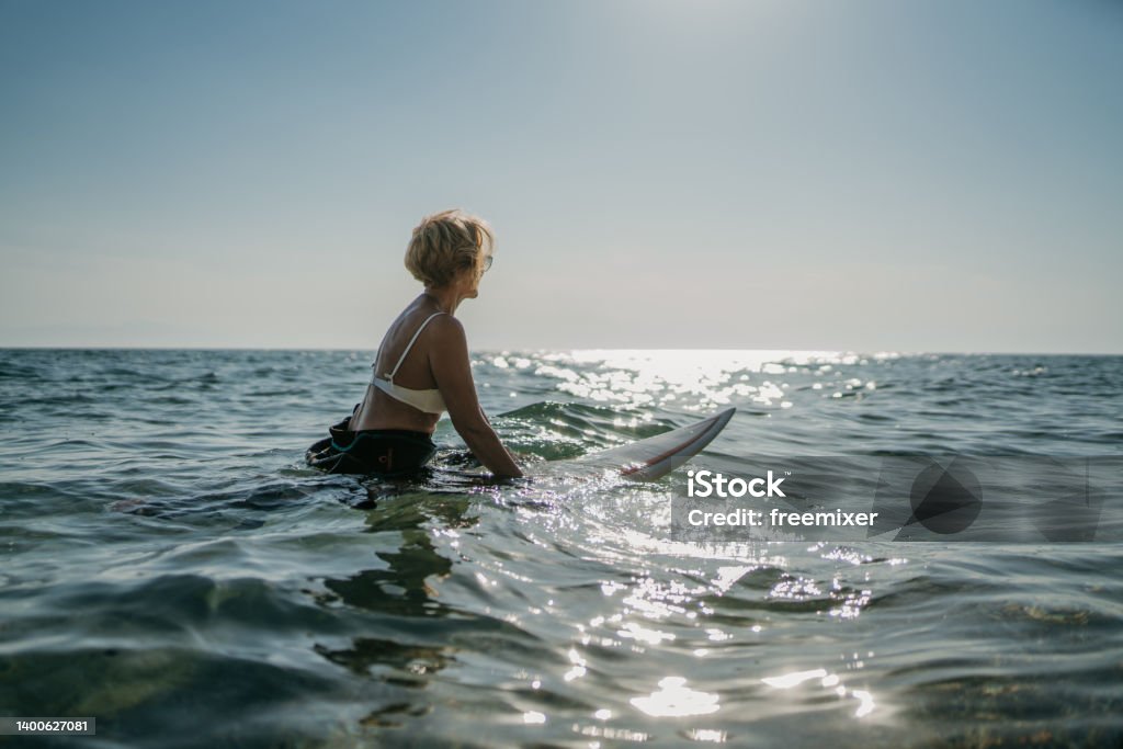 Female surfer in the sea Mature woman surfer sitting on surfboard in the Aegean sea. Surfing Stock Photo