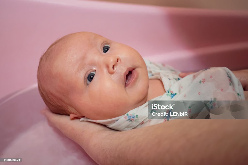 Mother washes newborn baby in pink bath with water at home Mother washes newborn baby girl holding in pink bath with water at home. Confused cute little daughter bathes wrapped in swaddle diaper close view Baby - Human Age Stock Photo