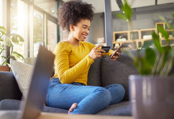 jeune femme métisse heureuse utilisant une carte de crédit et un téléphone seule à la maison. femme hispanique joyeuse avec un afro bouclé faisant un achat en ligne avec une carte de débit et un téléphone portable tout en étant assise sur le cana - livraison à domicile photos et images de collection